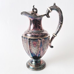 Op de afbeelding: Een vintage verzilverde kan met een gebogen handvat en een deksel. De kan heeft een sierlijk bloemmotief en een decoratief handvat met witte accenten. De kan is ongeveer 25 cm hoog.