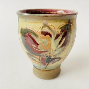 May include: A small, hand-thrown ceramic cup with a brown, yellow and red floral design. The cup has a rounded base and a flared rim.