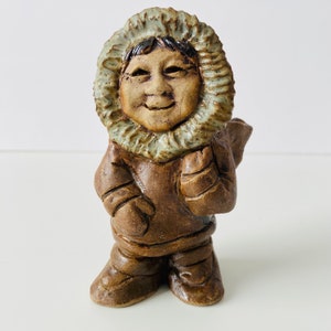 Könnte beinhalten: Eine Keramikfigur eines Menschen, der einen braunen Kapuzenparka mit weißem Pelzbesatz trägt. Die Person hat ein lächelndes Gesicht mit braunen Augen und einem braunen Bart.