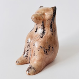 Könnte beinhalten: Eine sitzende Bärenfigur aus Keramik mit einer glänzenden beigen Glasur und dunkelbraunen, gesprenkelten und abgenutzt wirkenden Akzenten auf der Oberfläche. Der Bär hat eine glatte, abgerundete Form mit spitzen Ohren und einer ausgeprägten Schnauze.