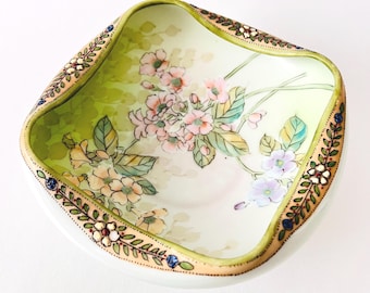 May include: A square, off-white ceramic bowl with a floral design. The interior features pink and yellow flowers with green leaves against a pale green background. The rim is decorated with a gold border and floral accents.