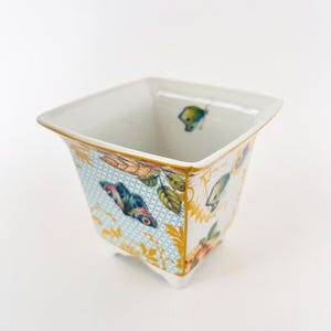 Könnte beinhalten: Quadratischer weißer Keramik-Pflanztopf mit einem dekorativen Schmetterlings- und Blumendesign. Der Pflanztopf hat einen hellblauen Hintergrund mit goldenen Akzenten und einen weißen Innenraum. Der Pflanztopf hat eine quadratische Form und wird von kleinen Füßen angehoben.