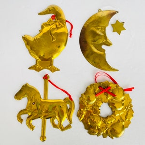 May include: Four brass-coloured ornaments: a duck, a crescent moon with a face, a star, a carousel horse, and a wreath. Each ornament has a red ribbon for hanging. The ornaments are on a white background.