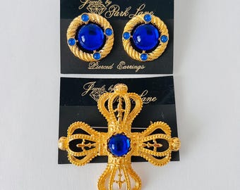 May include: A gold-coloured jewellery set comprising round pierced earrings and a cross-shaped brooch, each with a large, bright blue central stone. The earrings have a rope-like border. The brooch is crown-shaped.