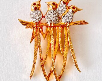 May include: A gold-toned brooch featuring four birds perched on a branch. The birds have clear crystal bodies and red eye accents. The brooch has a textured, detailed design.