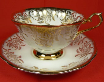 May include: A white teacup and saucer set with gold trim and a gold floral design. The cup has a gold handle and a scalloped rim. The saucer also has a scalloped edge and gold accents. The set is on a red background.