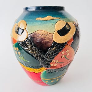 May include: A hand-painted ceramic vase with a detailed relief design. The vase features a dark blue rim, a landscape scene with figures wearing hats, and a colorful floral and geometric pattern. The vase is approximately 25 cm tall.