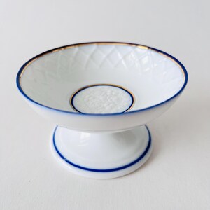 May include: A white porcelain pedestal dish with a blue and gold rim. The dish has a raised, textured centre.