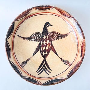 May include: A round, cream-colored ceramic plate with a hand-painted bird design. The bird has outstretched wings and a diamond-patterned body. The rim features a repeating pattern in brown and red, with a red inner border.