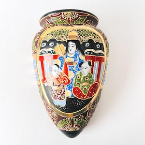 May include: A decorative, vintage Japanese vase with a heart-shaped design. The vase features a detailed illustration of figures in traditional attire, framed by gold accents and intricate floral patterns. The color palette includes red, blue, green, and gold.