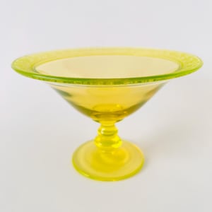 Peut inclure: Un bol sur pied en verre jaune translucide doté d'un bassin large et peu profond avec un bord texturé et à motifs. Le bol est soutenu par une tige courte à plusieurs niveaux qui se connecte à une base circulaire, créant une silhouette surélevée.