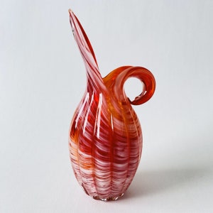 May include: A red and white glass vase with a swirl design. The vase has a handle and a rounded base.