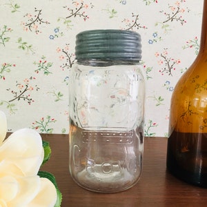 Peut inclure: Un bocal en verre transparent avec un couvercle en métal gris. Le bocal porte l'inscription "Crown" en relief sur le côté. Le bocal est posé sur une surface en bois.