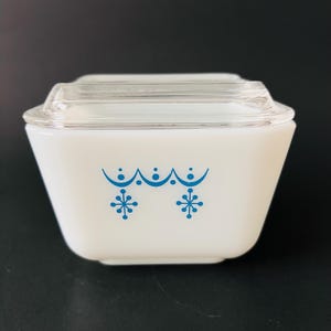 Op de afbeelding: Een vierkante voorraadbak van wit melkglas met een doorzichtig glazen deksel. De buitenzijde is voorzien van een blauw geprint patroon bestaande uit een geschulpte lijn met stippen en twee symmetrische stervormige motieven op de voorzijde.
