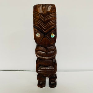 May include: A dark brown wooden tiki statue with a carved face and inlaid abalone shell eyes. The statue is standing on a white surface.