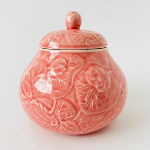 May include: A coral-coloured ceramic jar with a lid and a small knob on top. The jar and lid are decorated with a raised floral and leaf pattern. The jar has a round shape.