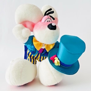 Könnte beinhalten: Ein Plüschtier des Weißen Kaninchens aus Alice im Wunderland, das eine blaue Jacke mit gelb-blau gestreiften Hosen, eine gelbe Fliege und einen blauen Zylinder mit floralen Akzenten trägt. Das Kaninchen hat rosa Ohren und ein rosa Gesicht.