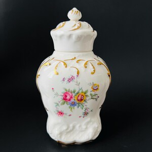 May include: A white porcelain jar with a lid, decorated with gold detailing and a floral pattern of pink, yellow, and blue flowers. The jar has a rounded shape and an ornate lid.