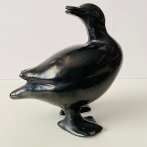 May include: Black ceramic sculpture of a duck with a long neck and a rounded body. The duck is standing on two legs with webbed feet.