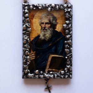 Pode incluir: Um ícone religioso emoldurado em prata com uma imagem de um homem com barba branca e um livro. O ícone está pendurado em uma corrente de contas de cor âmbar.