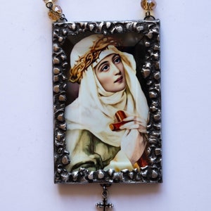 Puede incluir: Una imagen enmarcada en plata de una mujer con un velo blanco y una corona de espinas. La imagen cuelga de una cadena de cuentas con cuentas verdes y doradas. Una cruz de plata cuelga de la parte inferior del marco.