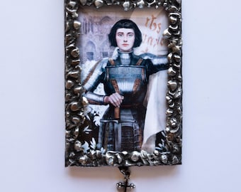 Könnte beinhalten: Ein silberfarbener Rahmen mit einer schwarzen Perlenkette, der ein Porträt von Jeanne d'Arc in Rüstung zeigt. Das Bild ist von einem weißen Rand umgeben und der Text "this mayle" ist in der oberen rechten Ecke sichtbar. Ein silbernes Kreuz hängt am unteren Rand des Rahmens.