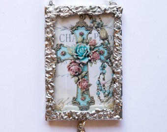 May include: A silver-framed ornament with a blue and white cross decorated with pink roses and a beaded rosary. The ornament has a crystal bead hanging from the bottom and a silver chain for hanging.