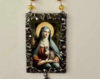 Könnte beinhalten: Ein silbernes gerahmtes Bild einer Frau in einem blauen Gewand, die ein Buch und eine Katze hält. Das Bild hängt an einer Kette mit goldenen Perlen. Ein silbernes Kreuz hängt unter dem Bild.