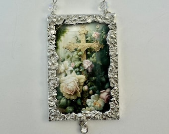 May include: A silver framed picture with a floral design and a cross in the center. The picture is hanging from a crystal beaded chain.