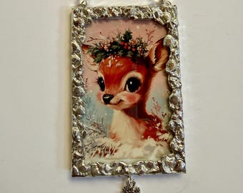 Puede incluir: Adorno navideño rectangular con una ilustración vintage de un ciervo con una corona floral. La imagen está enmarcada en un marco plateado, con un asa de cuentas y un dije de copo de nieve. El ciervo tiene pelaje marrón y ojos grandes y oscuros.