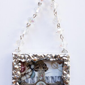 May include: A silver framed picture with a cross and flowers inside. The frame has a beaded chain attached to the top for hanging.