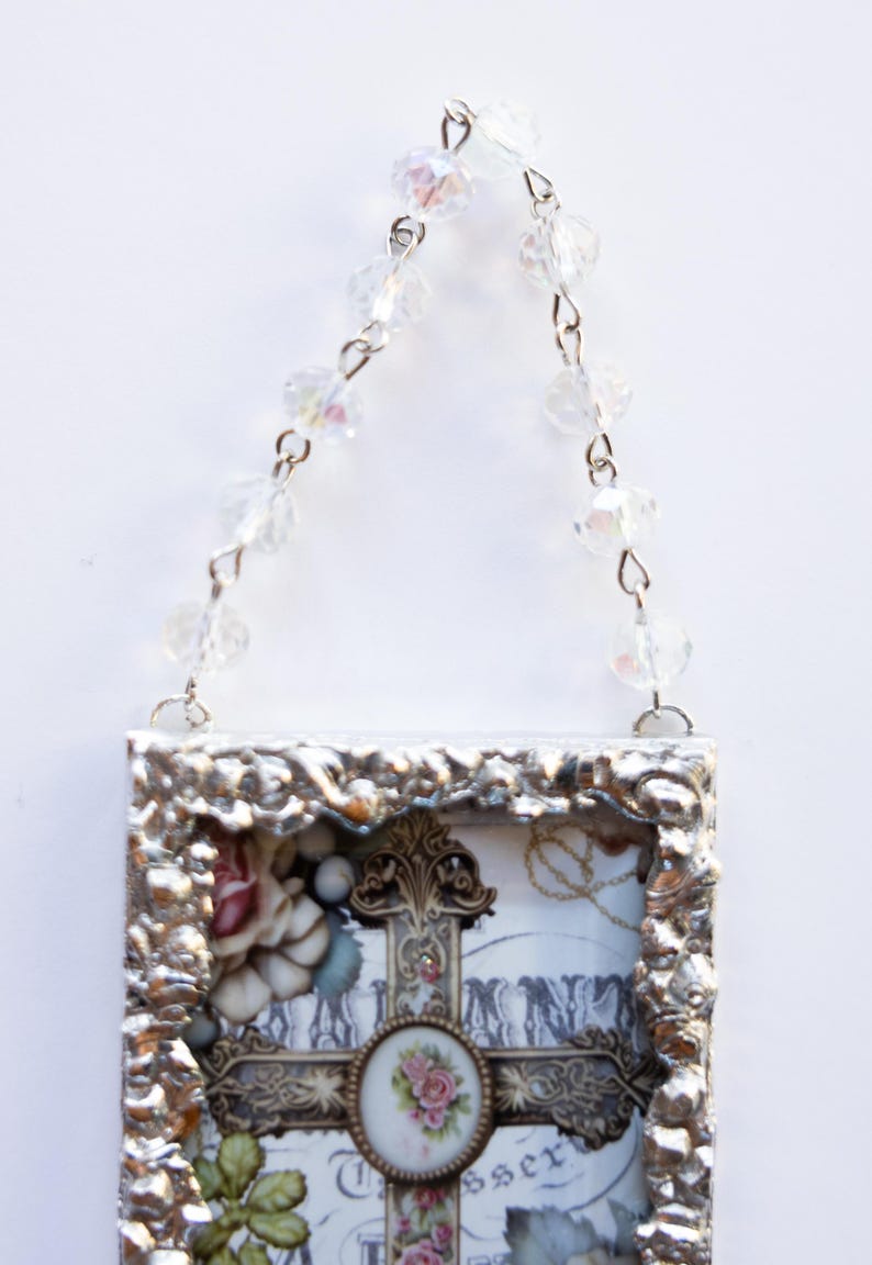 May include: A silver framed picture with a cross and flowers inside. The frame has a beaded chain attached to the top for hanging.