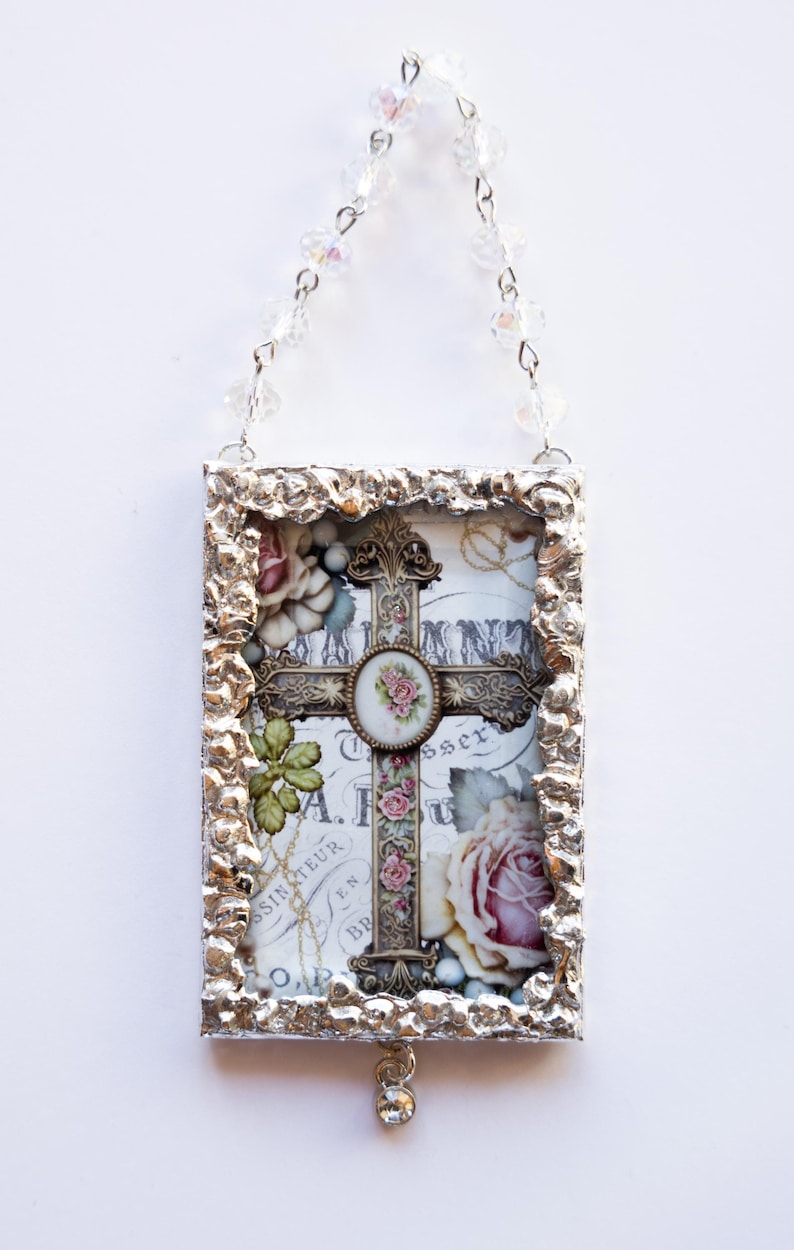 May include: A silver framed ornament with a cross and floral design. The cross is made of metal and has a floral design around it. The ornament is hanging from a silver chain.