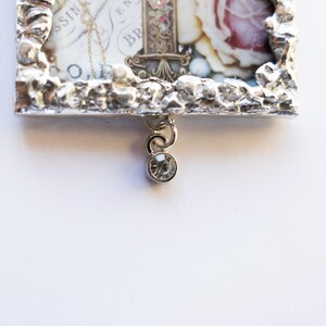 May include: A silver-toned picture frame with a textured border, featuring a vintage floral print with a rose and text. A small crystal charm hangs from the bottom of the frame.