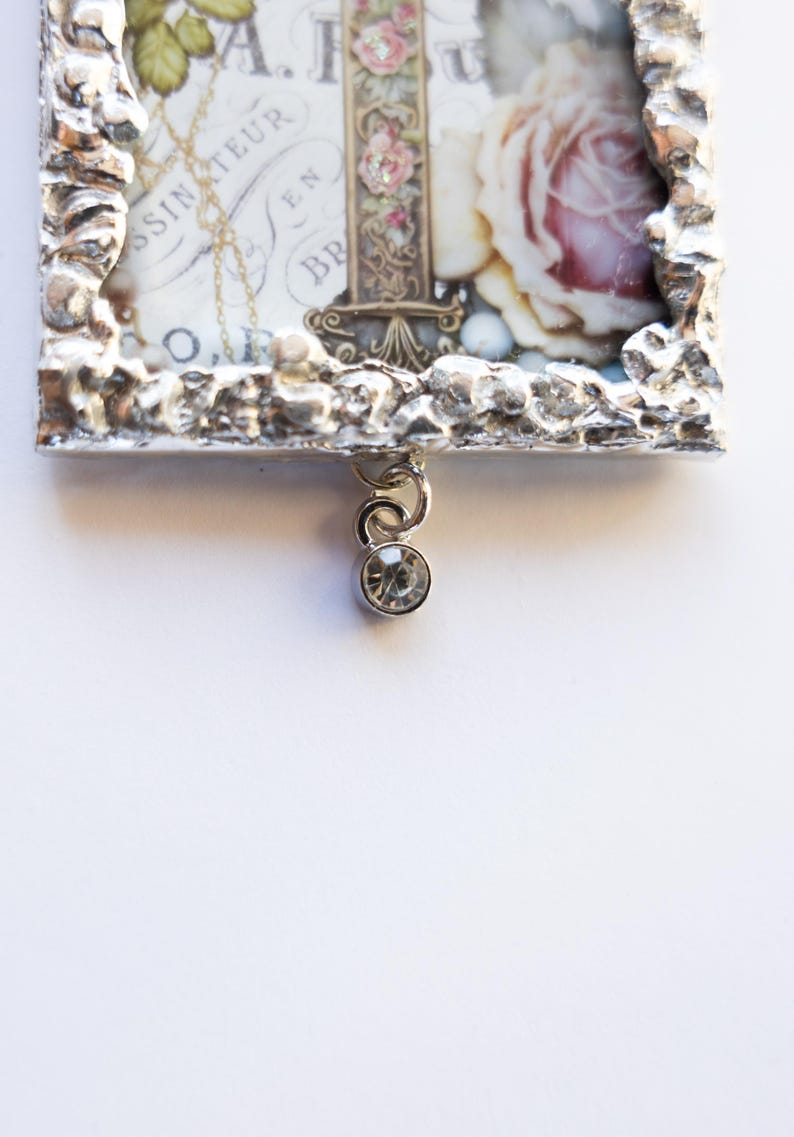 May include: A silver-toned picture frame with a textured border, featuring a vintage floral print with a rose and text. A small crystal charm hangs from the bottom of the frame.