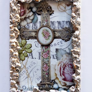 May include: A silver framed cross with a white background and floral accents. The cross is decorated with gold and has a small round floral design in the center. The frame has a textured, hammered metal look.