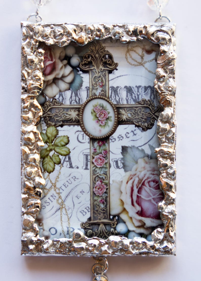 May include: A silver framed cross with a white background and floral accents. The cross is decorated with gold and has a small round floral design in the center. The frame has a textured, hammered metal look.