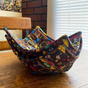 May include: A brown fabric bowl with a colorful print featuring characters and objects from the TV show "Friends". The bowl is folded in half and has a slightly curved shape.