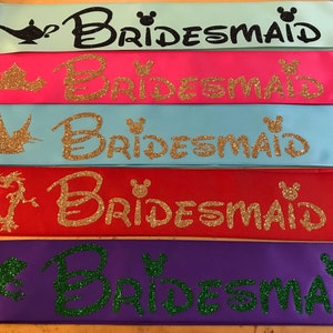 Puede incluir: Cinco cintas de sat&eacute;n en diferentes colores con la palabra "Bridemaid" escrita en purpurina y un personaje de Disney en cada cinta. Los personajes son una l&aacute;mpara, una corona, un p&aacute;jaro, un drag&oacute;n y una rana.