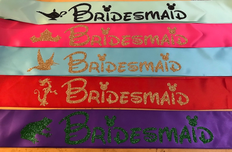 Puede incluir: Cinco cintas de sat&eacute;n en diferentes colores con la palabra "Bridemaid" escrita en purpurina y un personaje de Disney en cada cinta. Los personajes son una l&aacute;mpara, una corona, un p&aacute;jaro, un drag&oacute;n y una rana.
