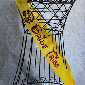 Puede incluir: Sash de sat&eacute;n amarillo con un dise&ntilde;o de purpurina rosa que dice "Bride Tribe" y presenta una rosa y una cabeza de Mickey Mouse de Disney.