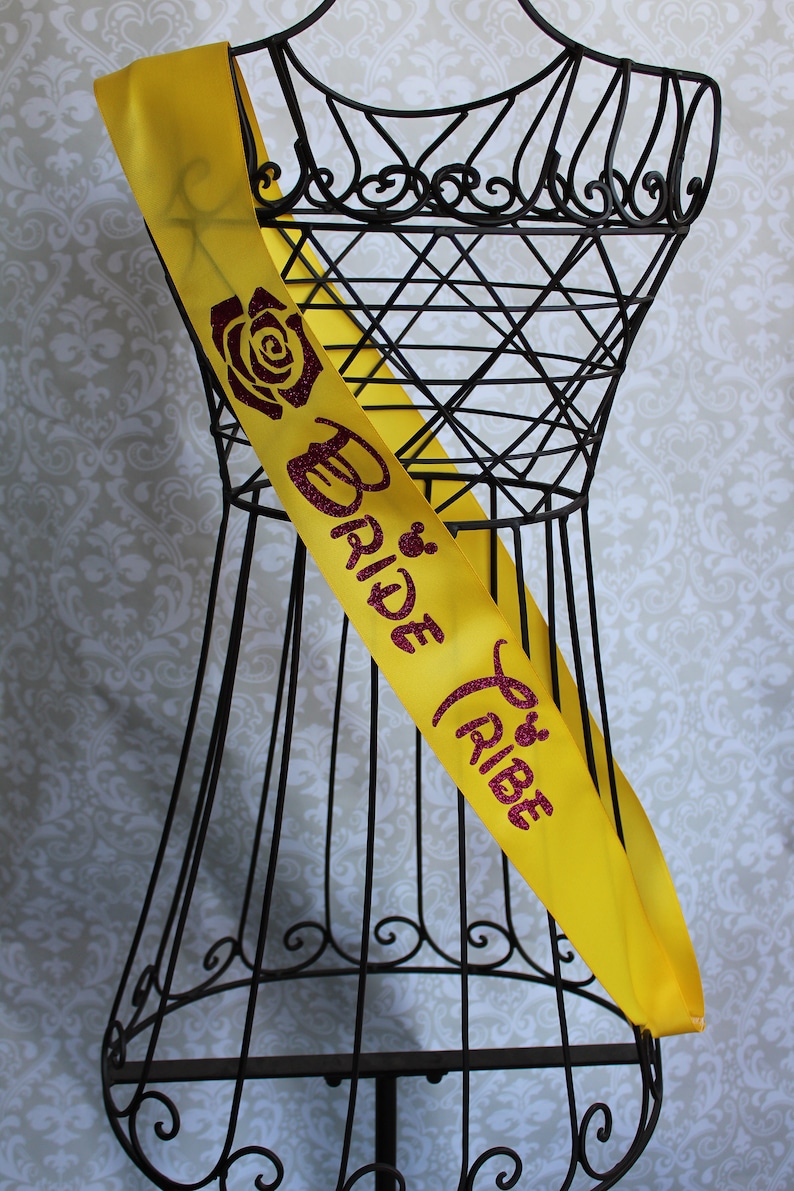 Puede incluir: Sash de sat&eacute;n amarillo con un dise&ntilde;o de purpurina rosa que dice "Bride Tribe" y presenta una rosa y una cabeza de Mickey Mouse de Disney.