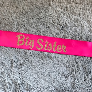 May include: Pink satin sash with gold glitter lettering that reads "Big Sister".