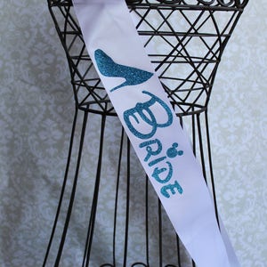 Puede incluir: Banda de sat&eacute;n blanco con un dise&ntilde;o de brillo azul de un zapato de tac&oacute;n alto y la palabra "Bride" con una peque&ntilde;a cabeza de Mickey Mouse.