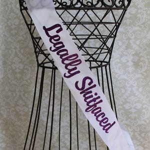 May include: White satin sash with the text "Legally Shitfaced" in purple glitter lettering.