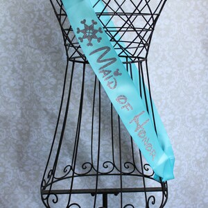 Puede incluir: Una banda de sat&eacute;n turquesa con letras plateadas de purpurina que dice "Maid of Honor" y un dise&ntilde;o de copo de nieve.