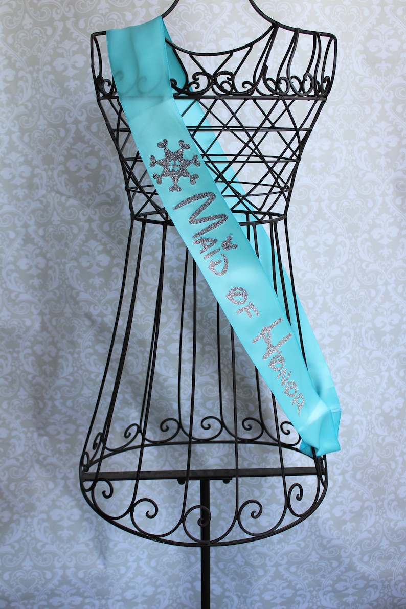 Puede incluir: Una banda de sat&eacute;n turquesa con letras plateadas de purpurina que dice "Maid of Honor" y un dise&ntilde;o de copo de nieve.