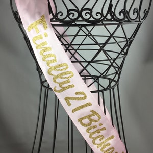 May include: Pink satin sash with gold glitter lettering that reads "Finally 21 Bitches!"