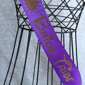 Puede incluir: Banda de sat&eacute;n morado con letras doradas de purpurina que dicen "Bride to Be" y un dise&ntilde;o de sol de purpurina dorada.