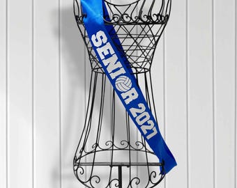 May include: A royal blue satin sash draped over a black wire dress form. The sash features the white text SENIOR 2021 with a volleyball graphic replacing the letter O. The fabric has a smooth, reflective finish and is worn diagonally across the torso.